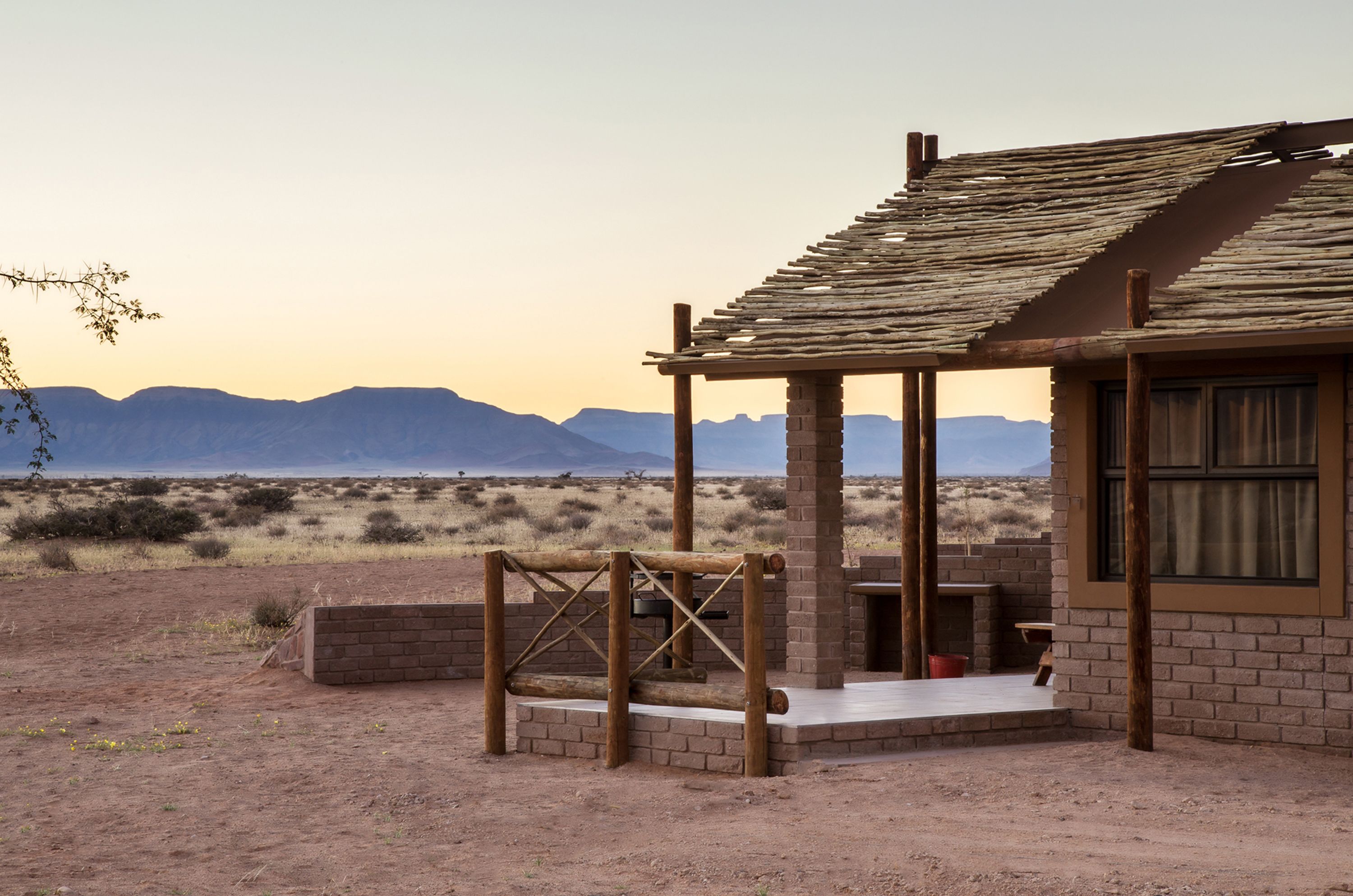 A rustic building with a brick exterior and a wooden roof, situated in a desert landscape with mountains in the background.