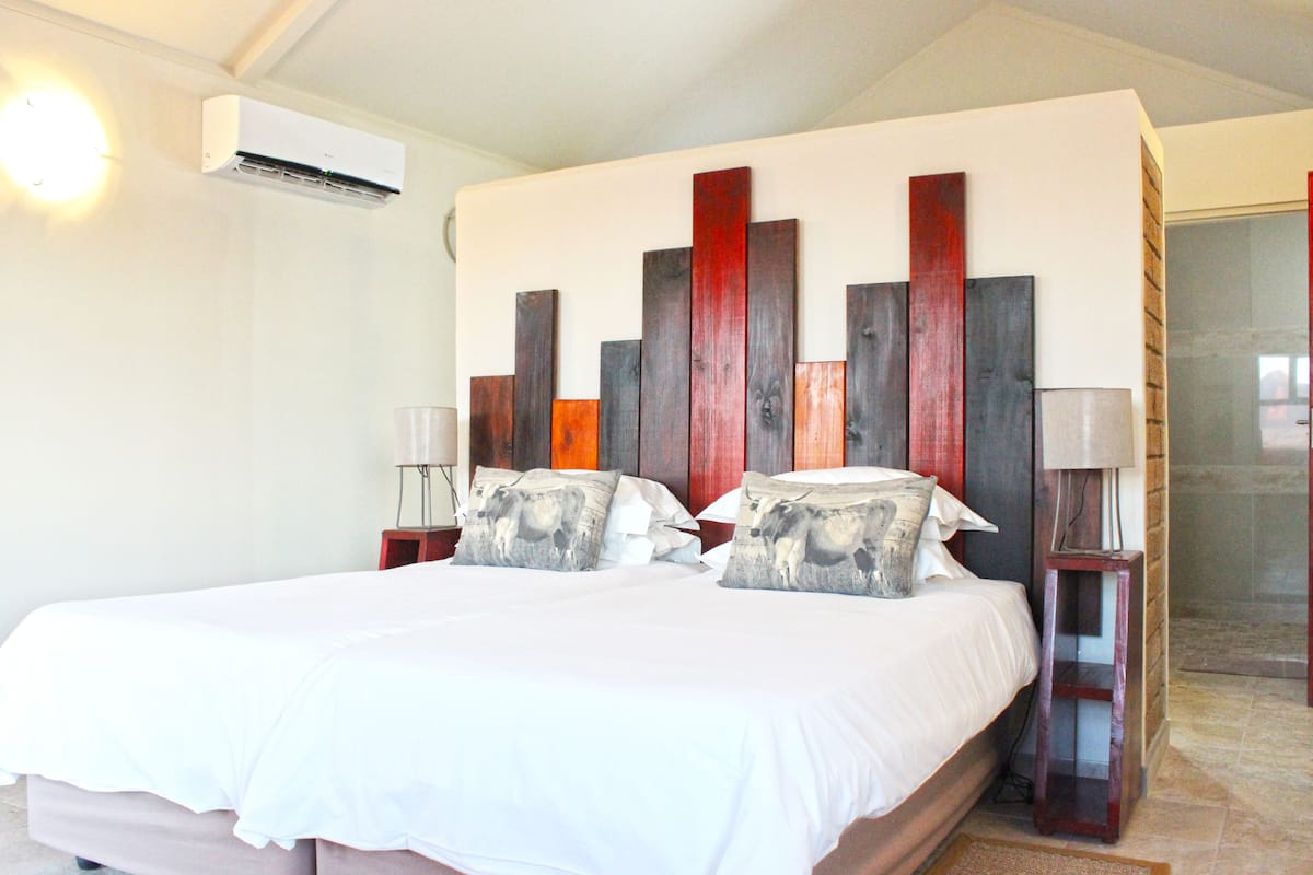 Uma cama de casal com colcha branca e duas almofadas decorativas, ar-condicionado fixado na parede e mesa de cabeceira com abajur.