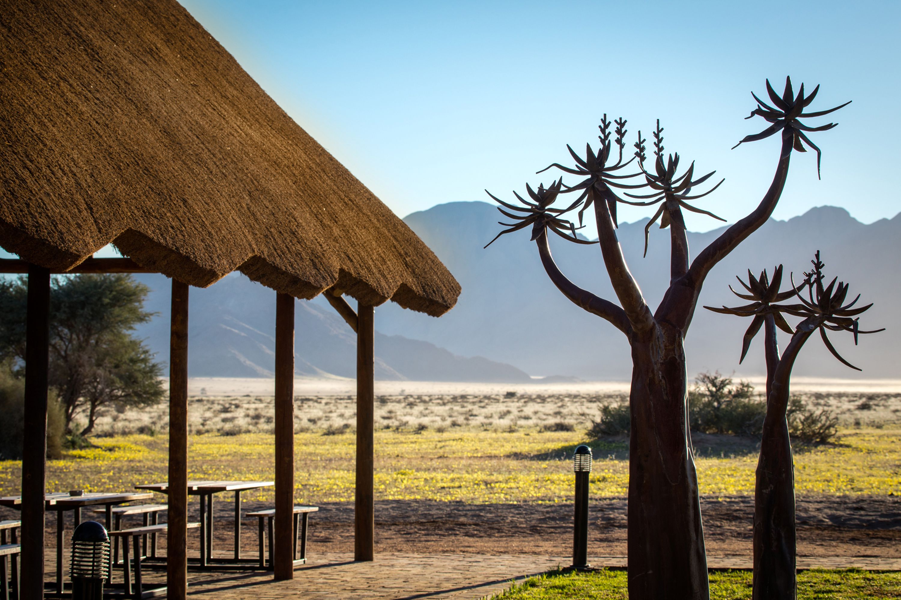 Una estructura con techo de paja, con asientos al aire libre, una escultura de árbol y un paisaje desértico con montañas al fondo.