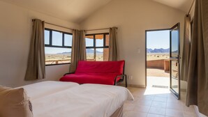 A bedroom with a bed, a red pillow, and a view of a desert landscape through a window. - Desert Camp (Sesriem)