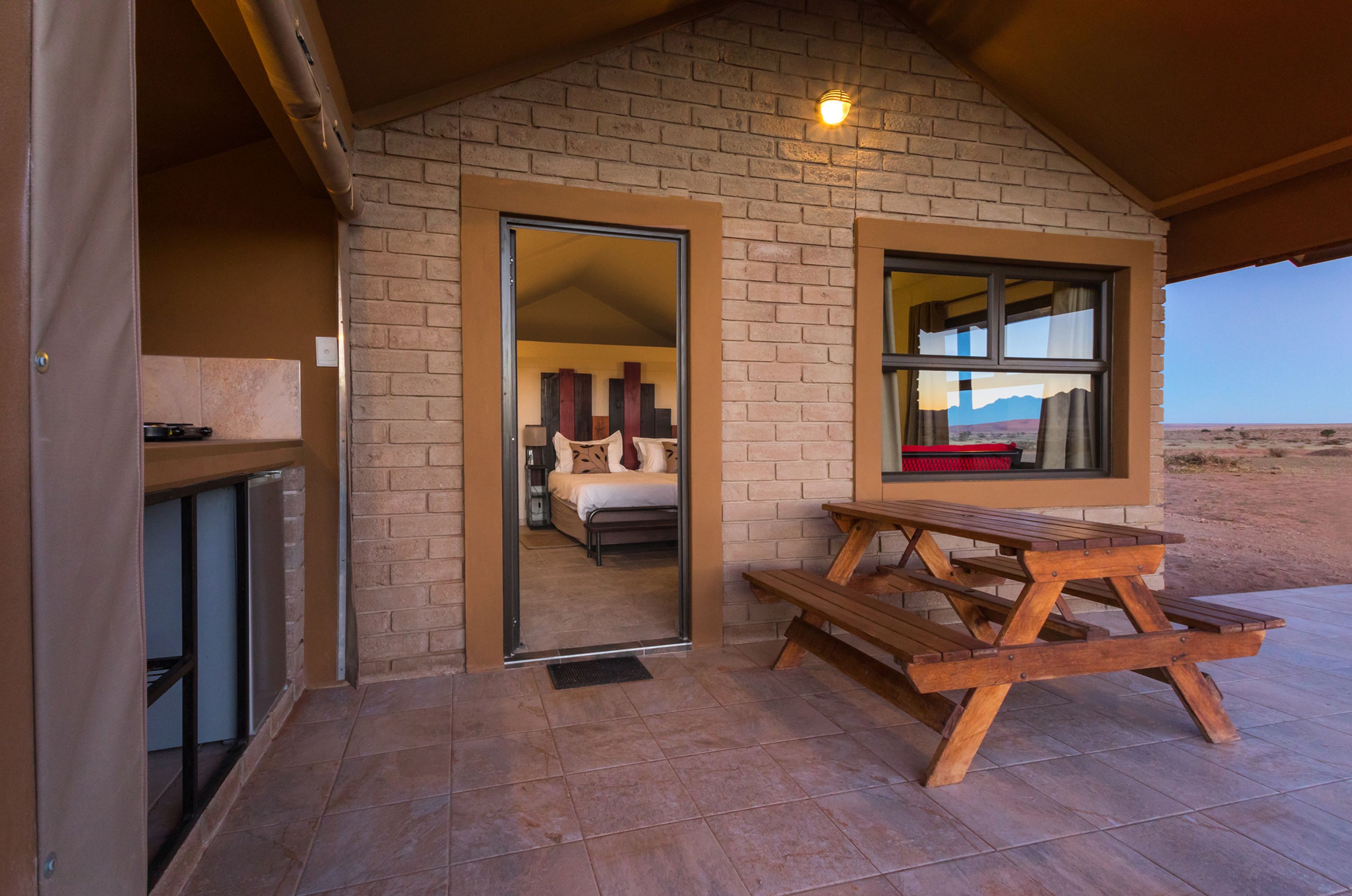 Eine rustikale Holzhütte mit Ziegelaußenwand, einer Holzbank und einem durchgehenden Türspalt einsehbaren Schlafzimmer.