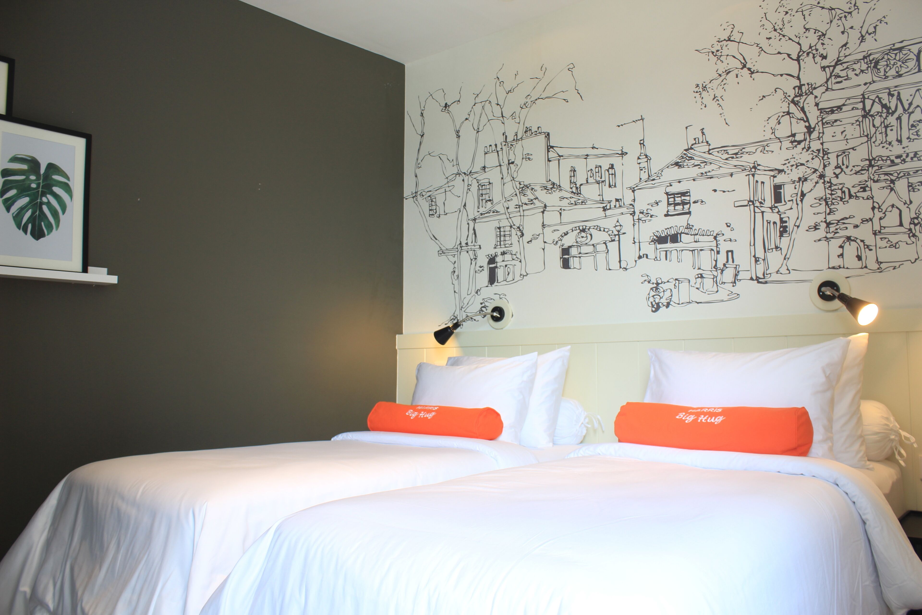 Une chambre d’hôtel avec un grand lit, du linge de lit blanc et un oreiller orange. Un mur est décoré d’une fresque représentant une scène de ville.