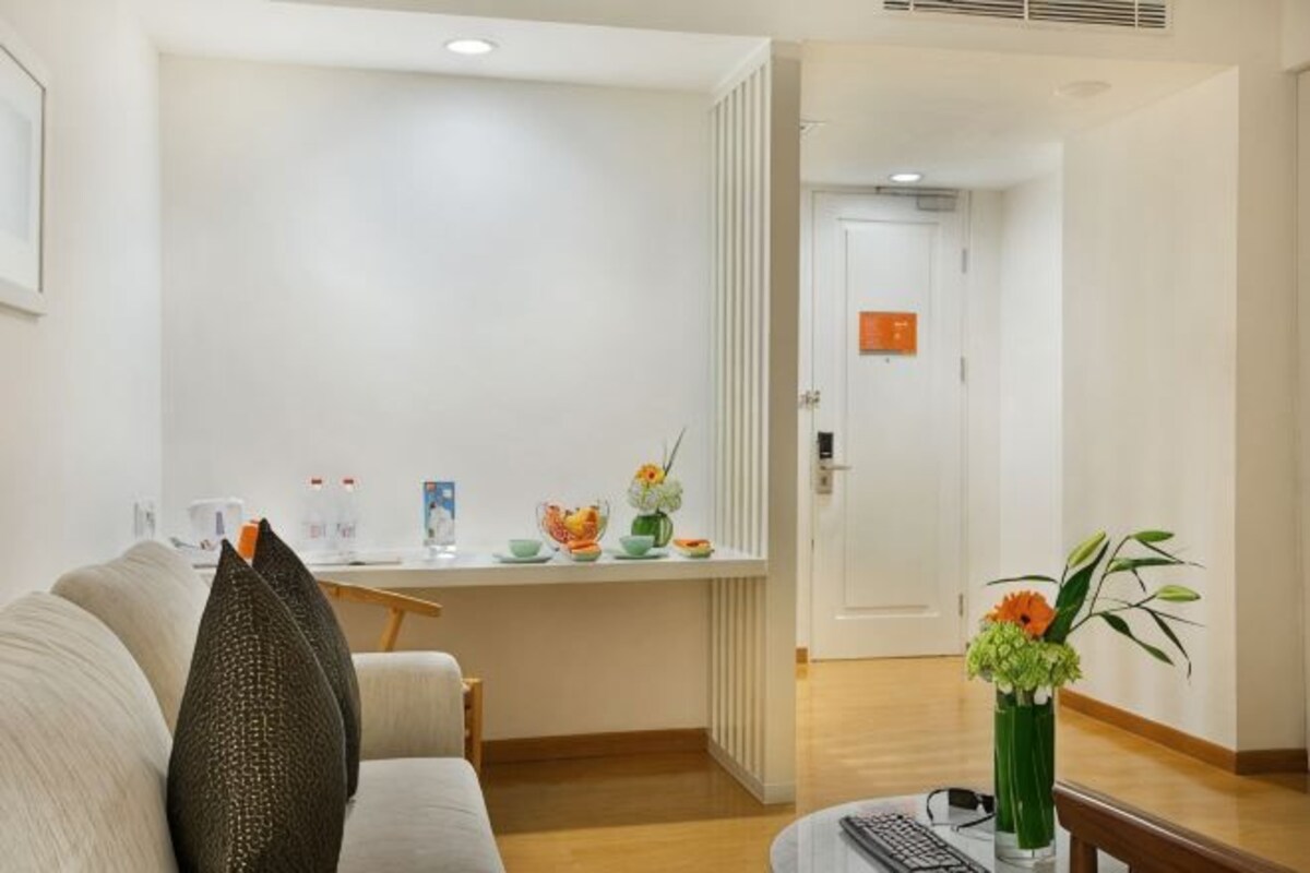Habitación de hotel con sofá, una mesita con un florero, un escritorio con ordenador y una puerta que da a otra habitación.