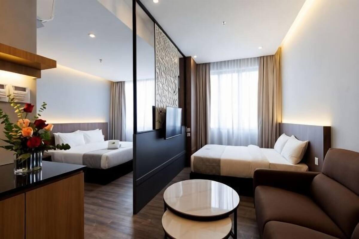 Habitación de hotel con dos camas, un sofá, un televisor, un escritorio con un florero y un ventanal con cortinas.