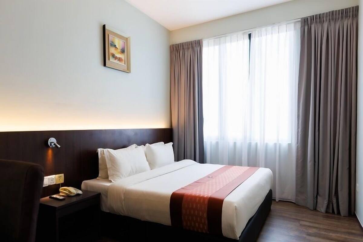 Una habitación de hotel con una cama grande, una mesita de noche con teléfono, una lámpara de pared y una ventana con cortinas.