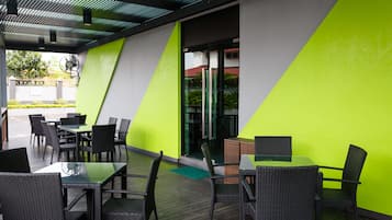 Un área de patio al aire libre con mesas y sillas, una pared verde y una puerta de vidrio que da al interior.