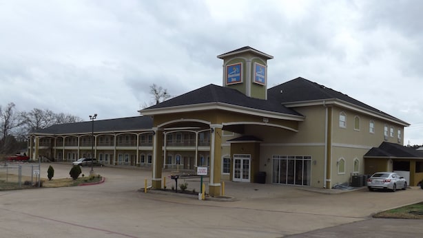 A large hotel with a central entrance, multiple parking spaces, and a covered waiting area. - SUNRISER INN (Kilgore)