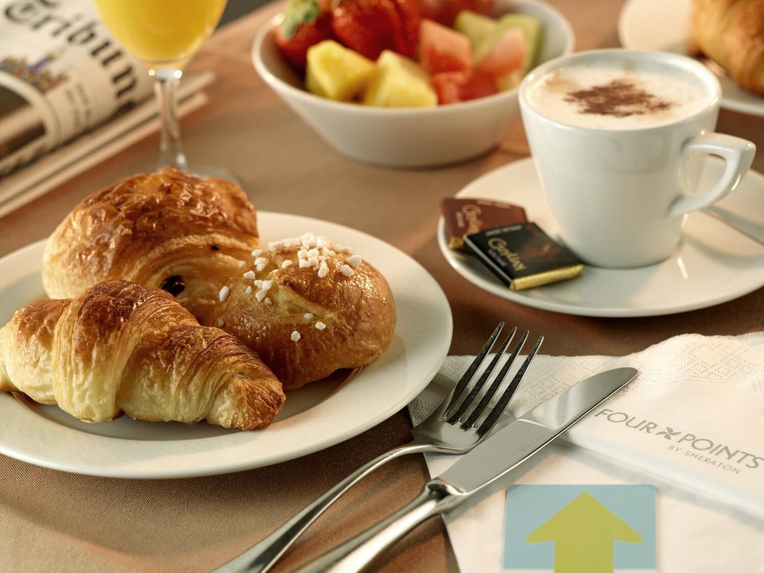 Een ontbijtsetting met croissants, een fruitschaal, een kop koffie en een krant.