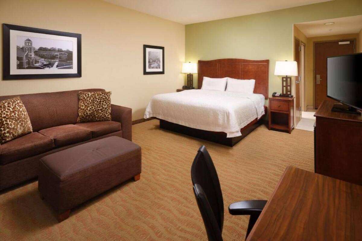 A hotel room with a bed, a sofa, a desk, a chair, a television, and framed pictures on the wall.