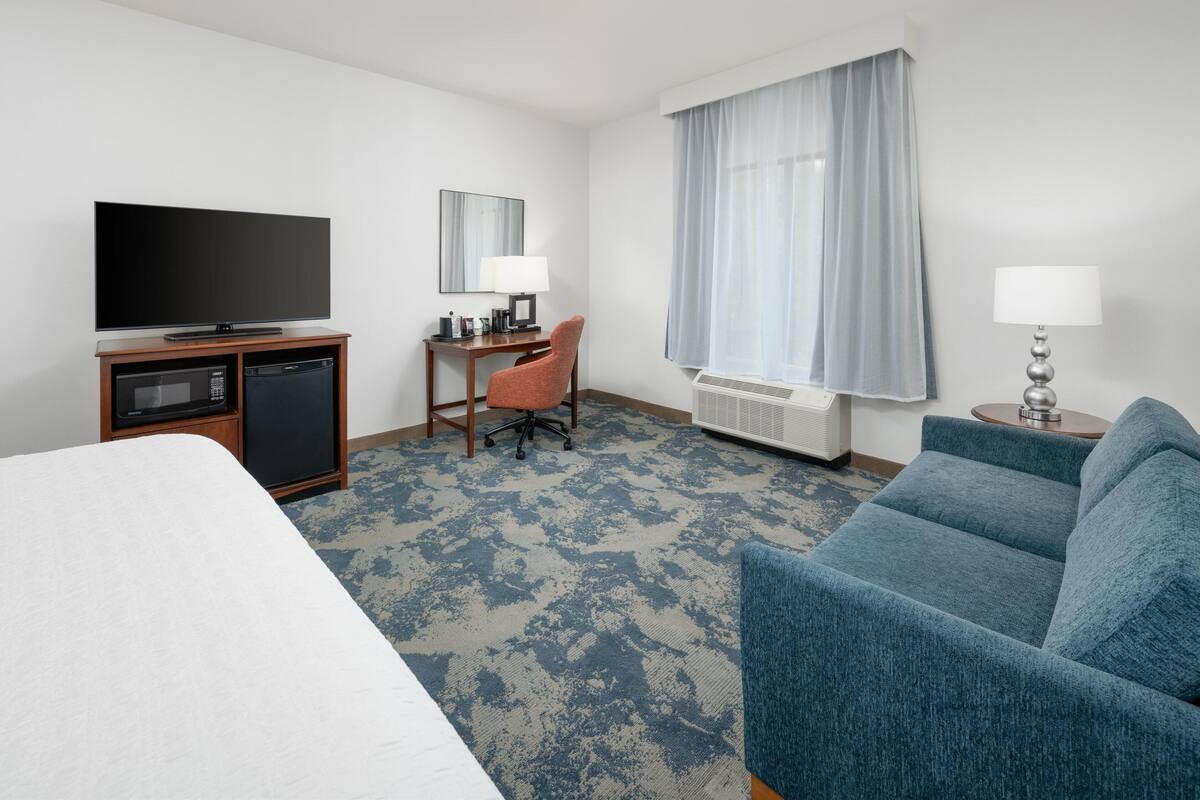 A hotel room with a bed, a TV, a desk with a chair, a blue sofa, and a window with curtains.