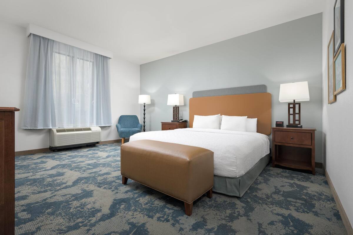 A hotel room with a large bed, a chair, two bedside tables with lamps, a window with curtains, and an air conditioning unit.