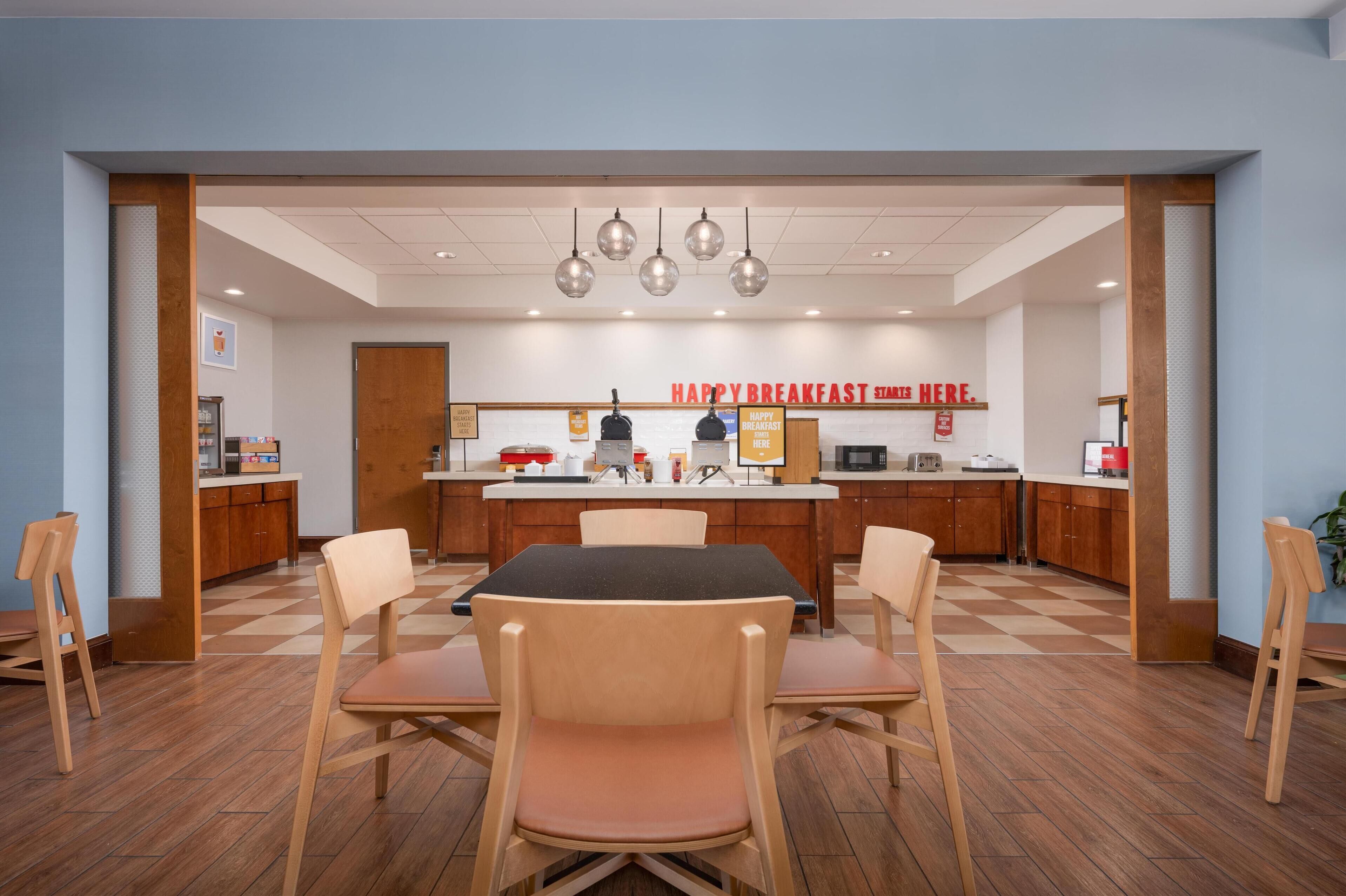 A breakfast area with a central table and chairs, a kitchen in the background, and a 'Happy Breakfast Here' sign.