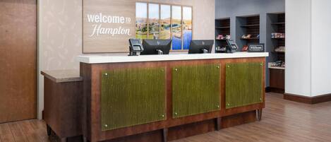 A hotel reception desk with a 'Welcome to Hampton' sign, a view of the landscape, and a 'Hilton Honors' sign.