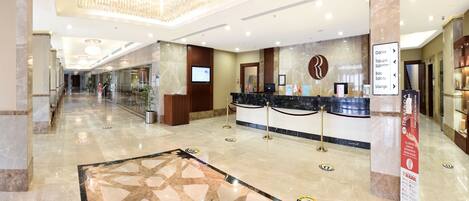 A hotel lobby with a reception desk, a digital screen, a marble floor with a star pattern, and a staircase in the background.