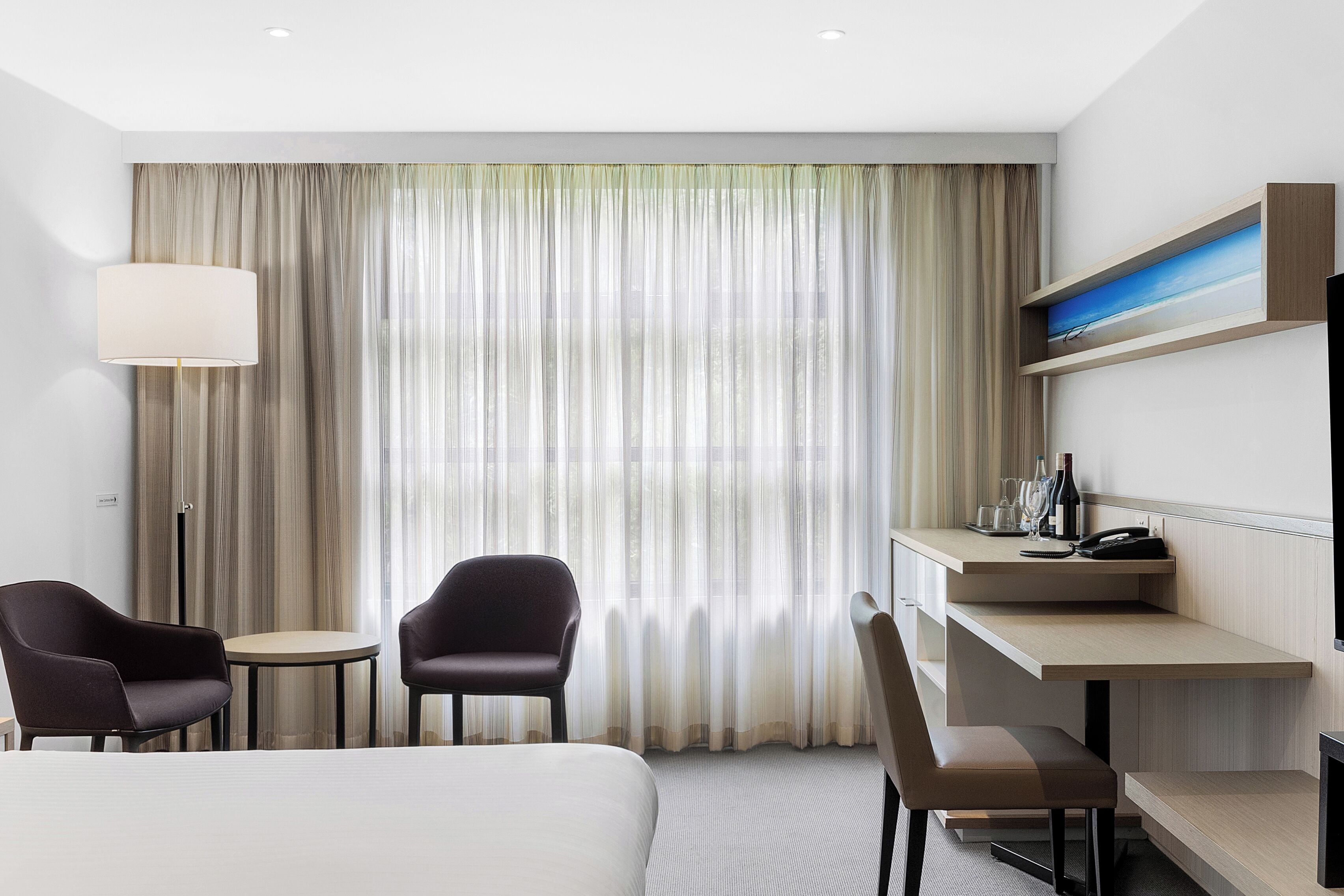 A hotel room with a bed, two armchairs, a desk, a lamp, a painting, and a large window with curtains.