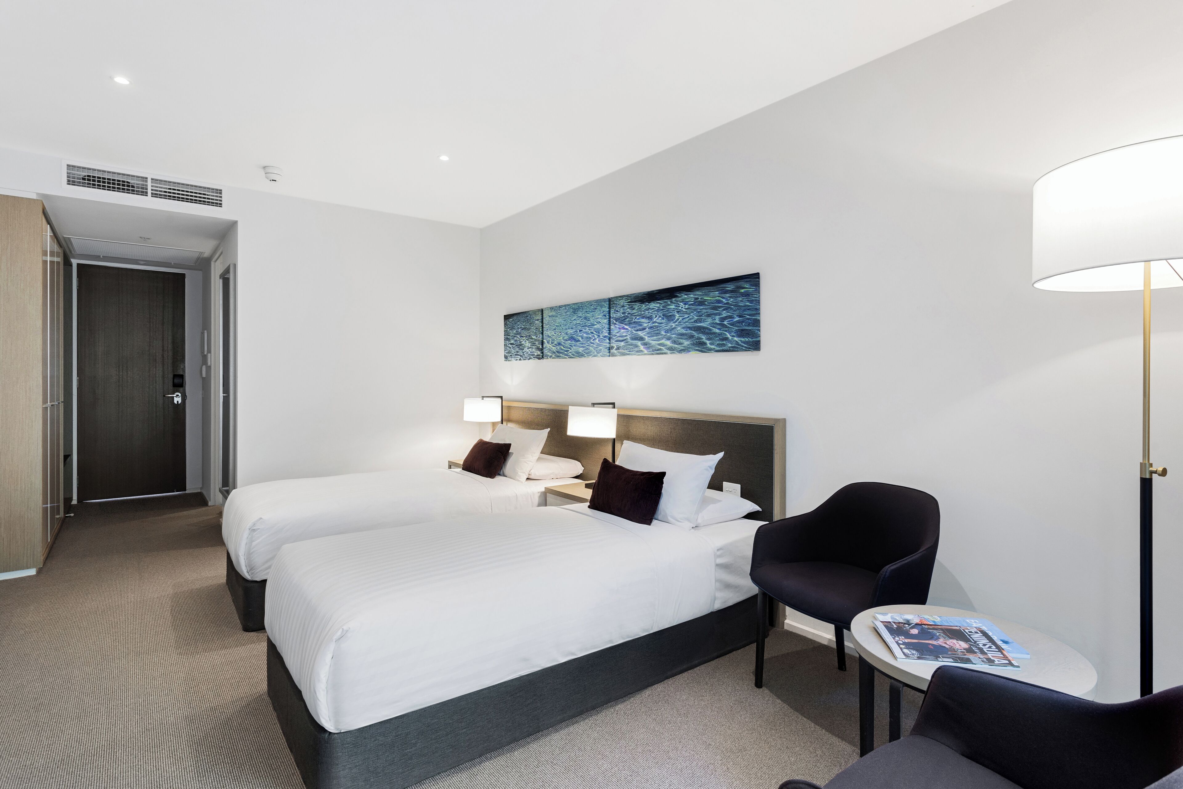 Quarto de hotel com duas camas, uma mesinha, uma cadeira e uma obra de arte na parede.