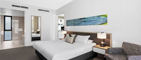 A modern hotel room with a large bed, a bedside table with a lamp, a patterned armchair, and a wall-mounted artwork.