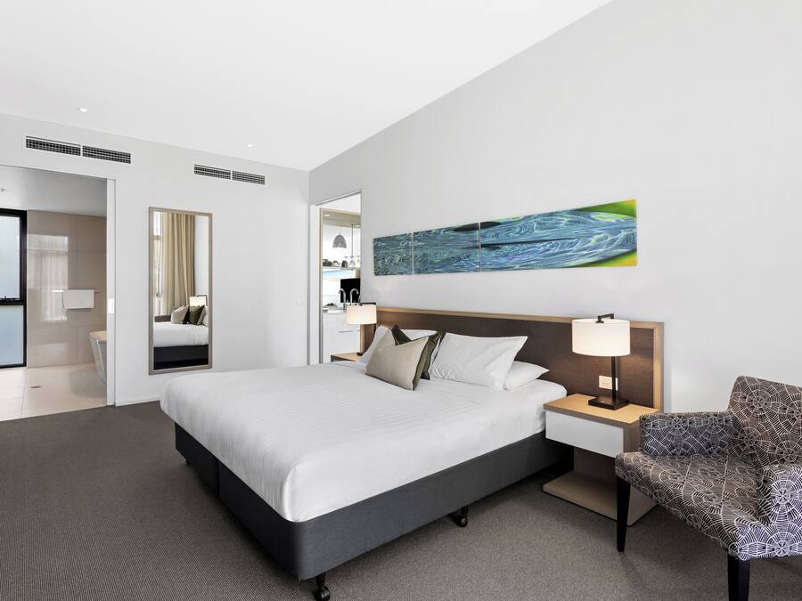 Una habitación de hotel moderna con una cama grande, una mesita de noche con lámpara, un sillón estampado y una obra de arte colgada en la pared.