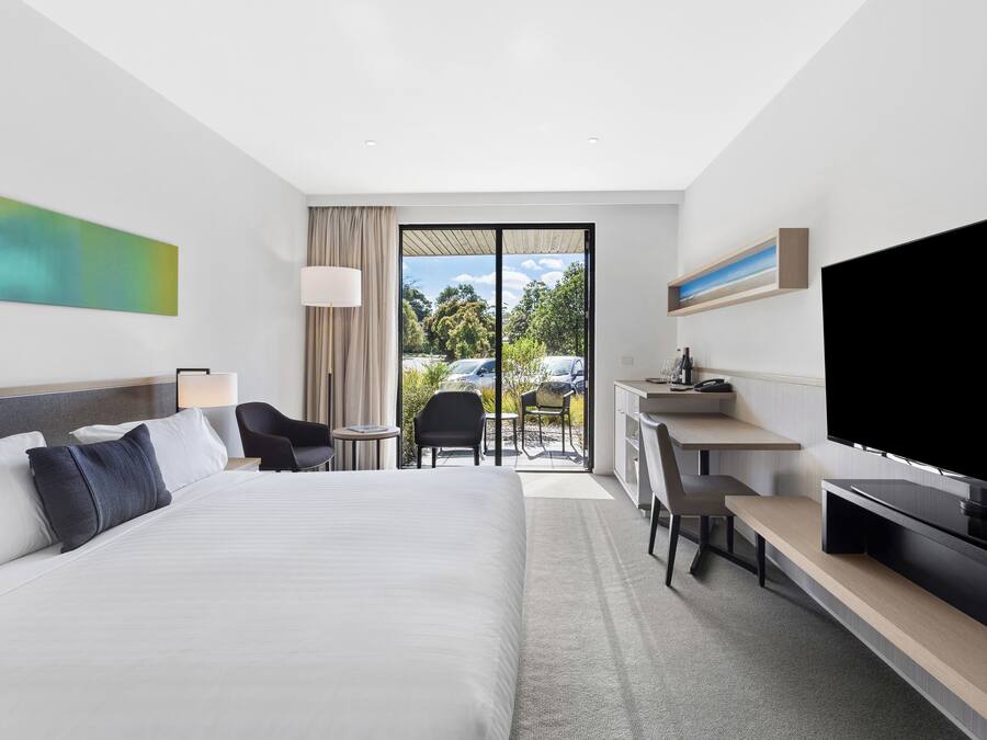 A modern hotel room with a large bed, a desk, a flat-screen TV, and a view of the outdoors.