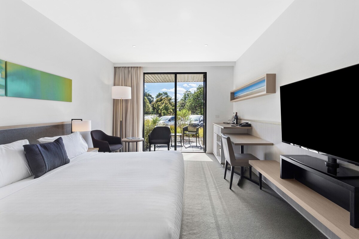 A modern hotel room with a large bed, a desk, a flat-screen TV, and a sliding glass door leading to an outdoor area.