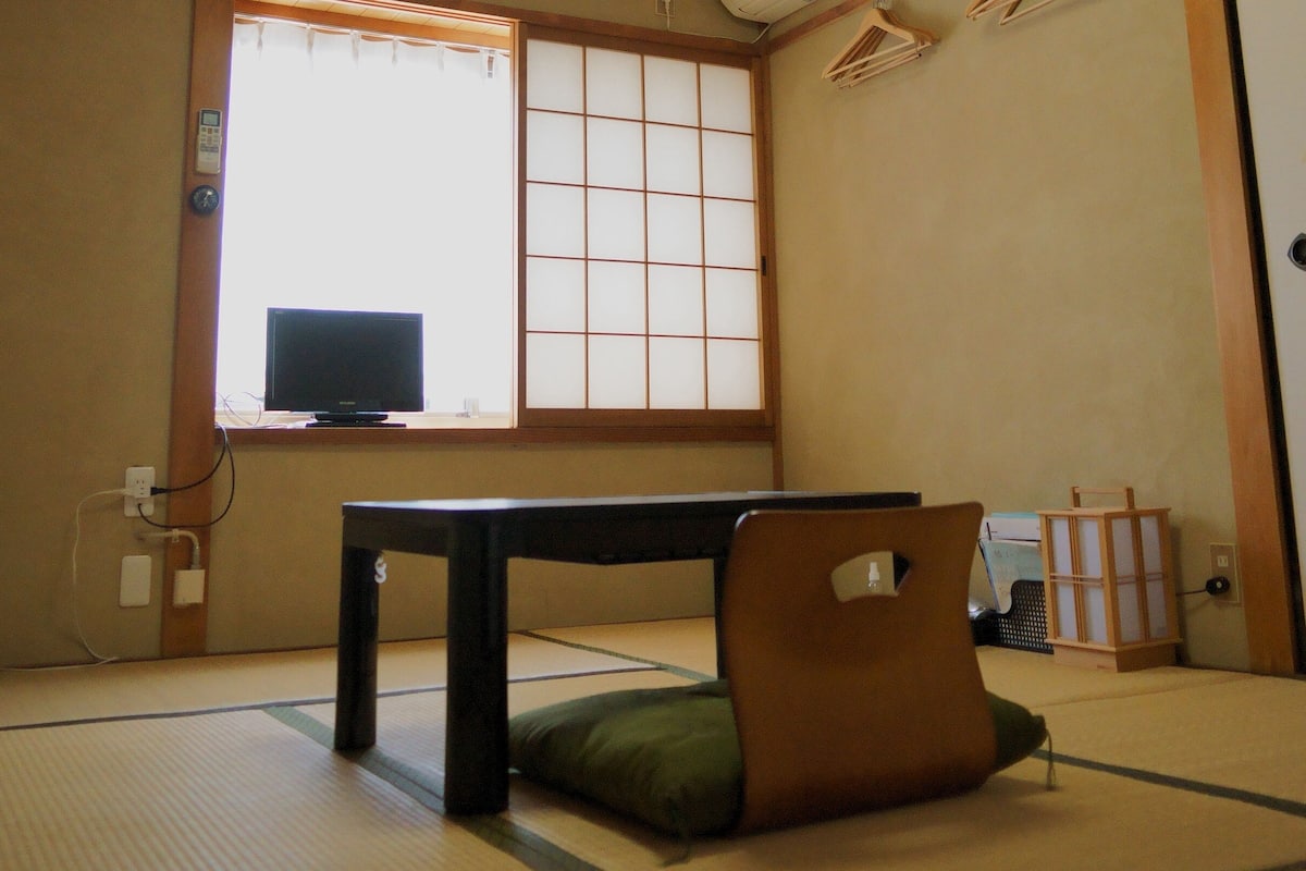 Quarto japonês tradicional com tatames, uma mesa baixa, um assento almofadado verde, um leque e uma televisão numa divisória deslizante.