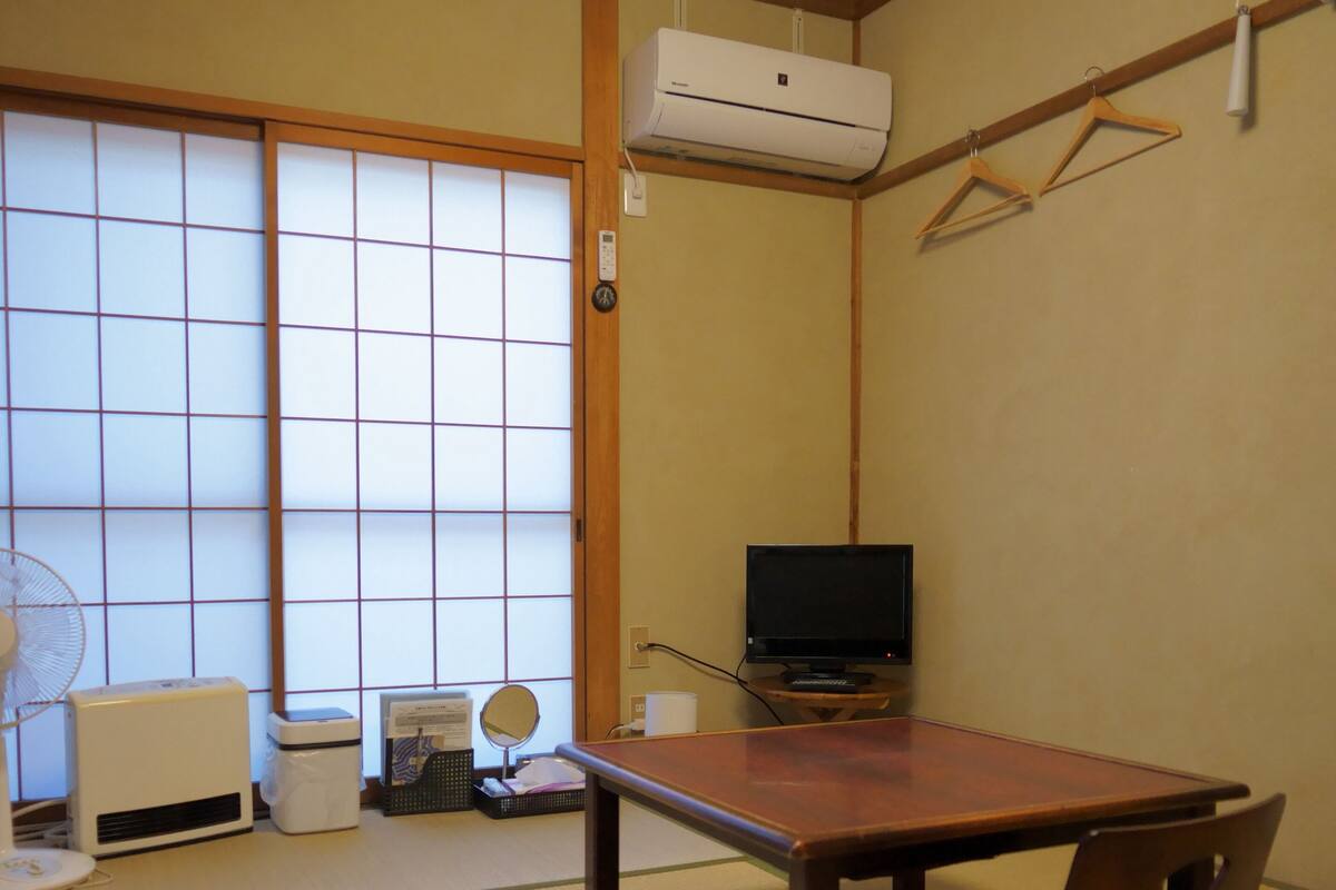 Um quarto japonês tradicional com piso de tatame, porta deslizante estilo shoji, ventoinha de teto, televisão e uma pequena mesa.