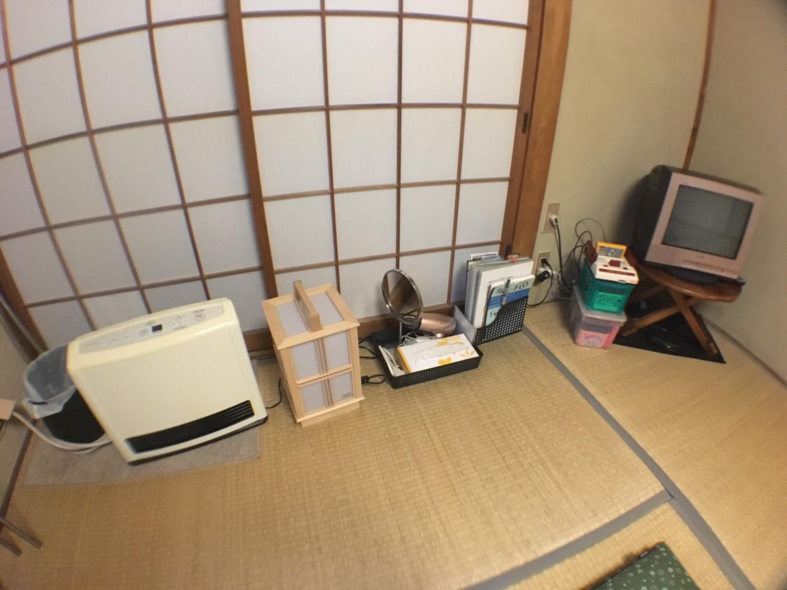 A room with tatami flooring, a traditional Japanese shoji screen, a white heater, a small wooden shelf, a desktop computer, and a CRT television.