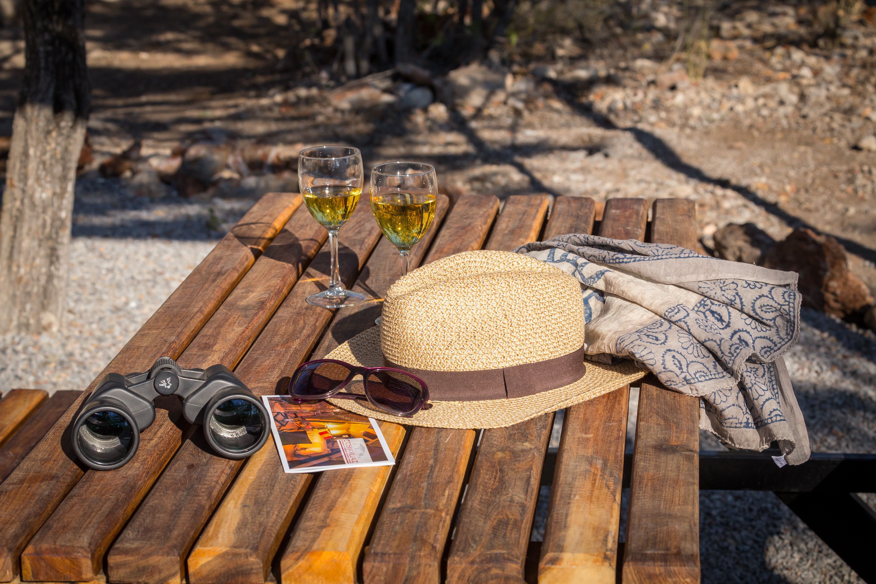 A wooden table with a straw hat, binoculars, sunglasses, and two glasses of wine.