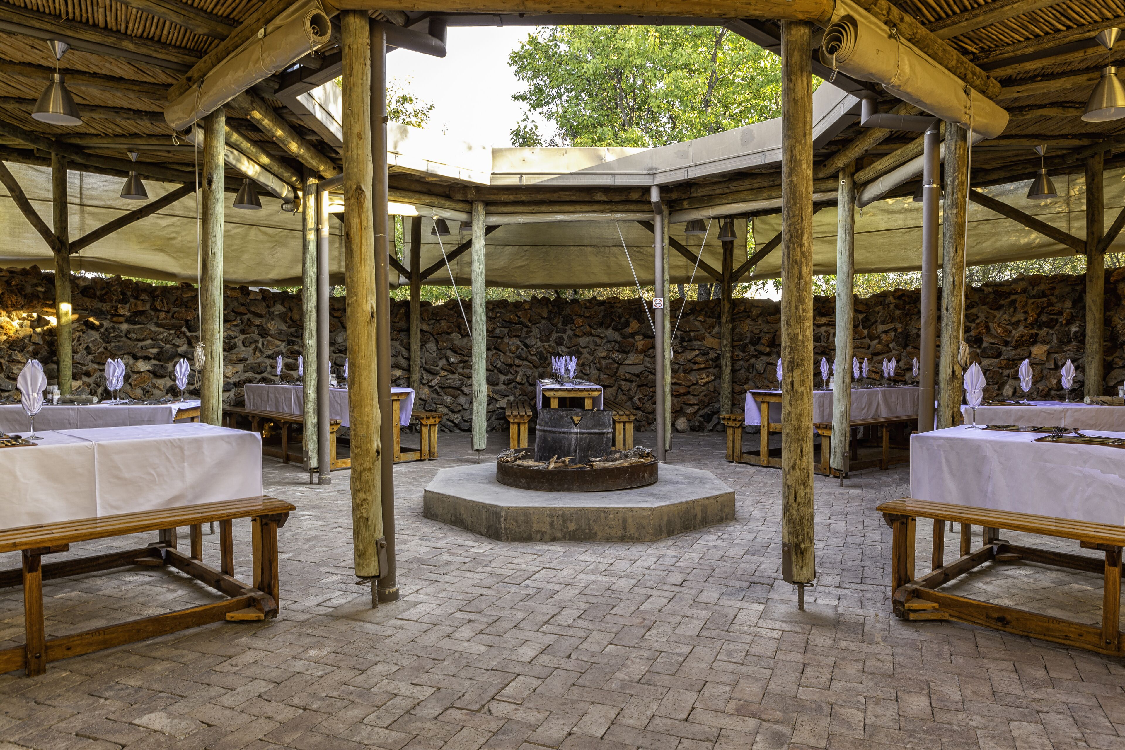 Un área de comedor al aire libre con techo, paredes de piedra, mesas de madera y bancos.