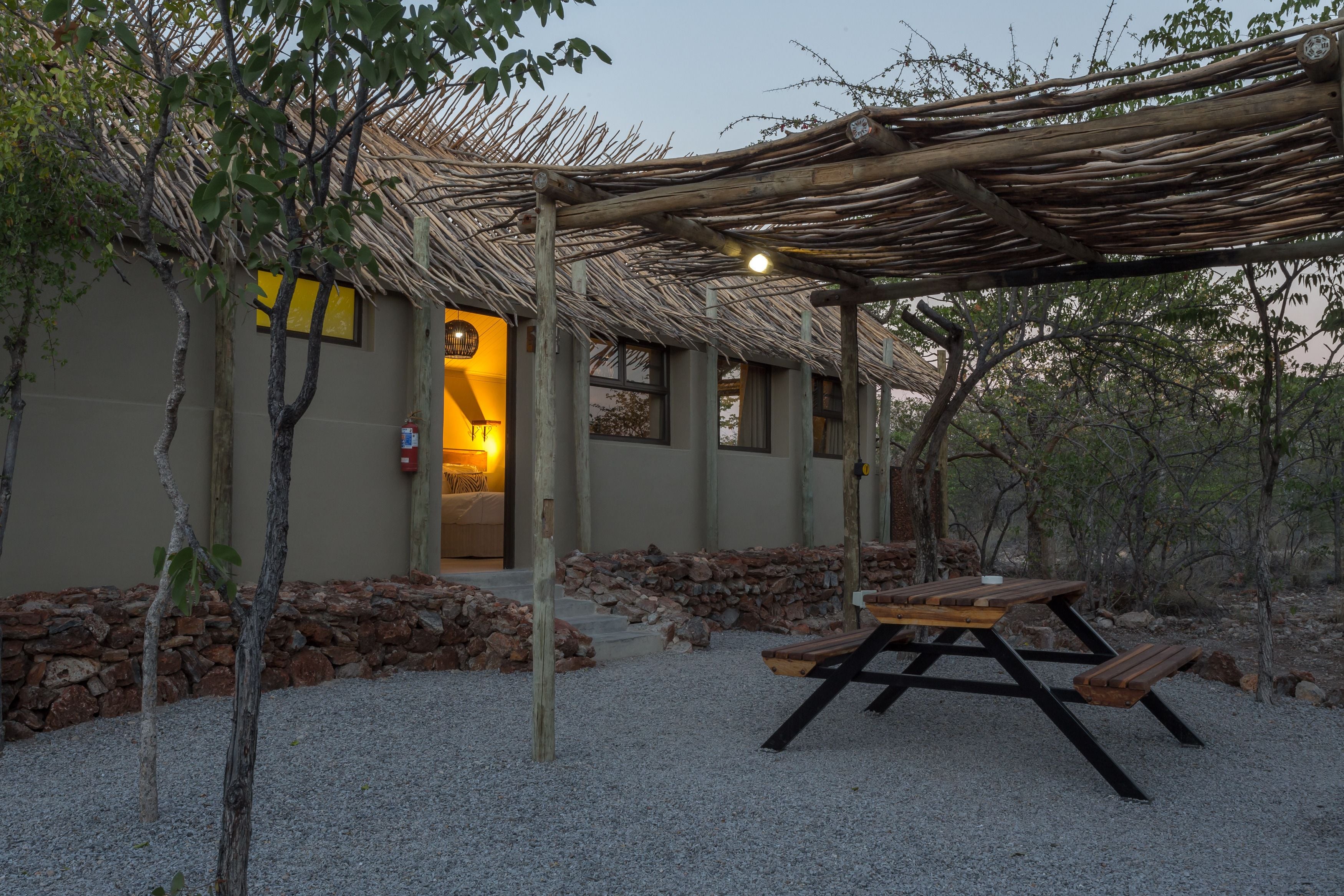 A rustic outdoor seating area with a wooden table and benches, surrounded by a gravel ground and a stone wall.