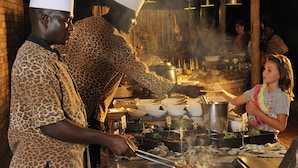 A chef in a leopard print uniform cooking at a busy outdoor market stall with various cooking utensils and ingredients visible. - Etosha Village (Okaukuejo)