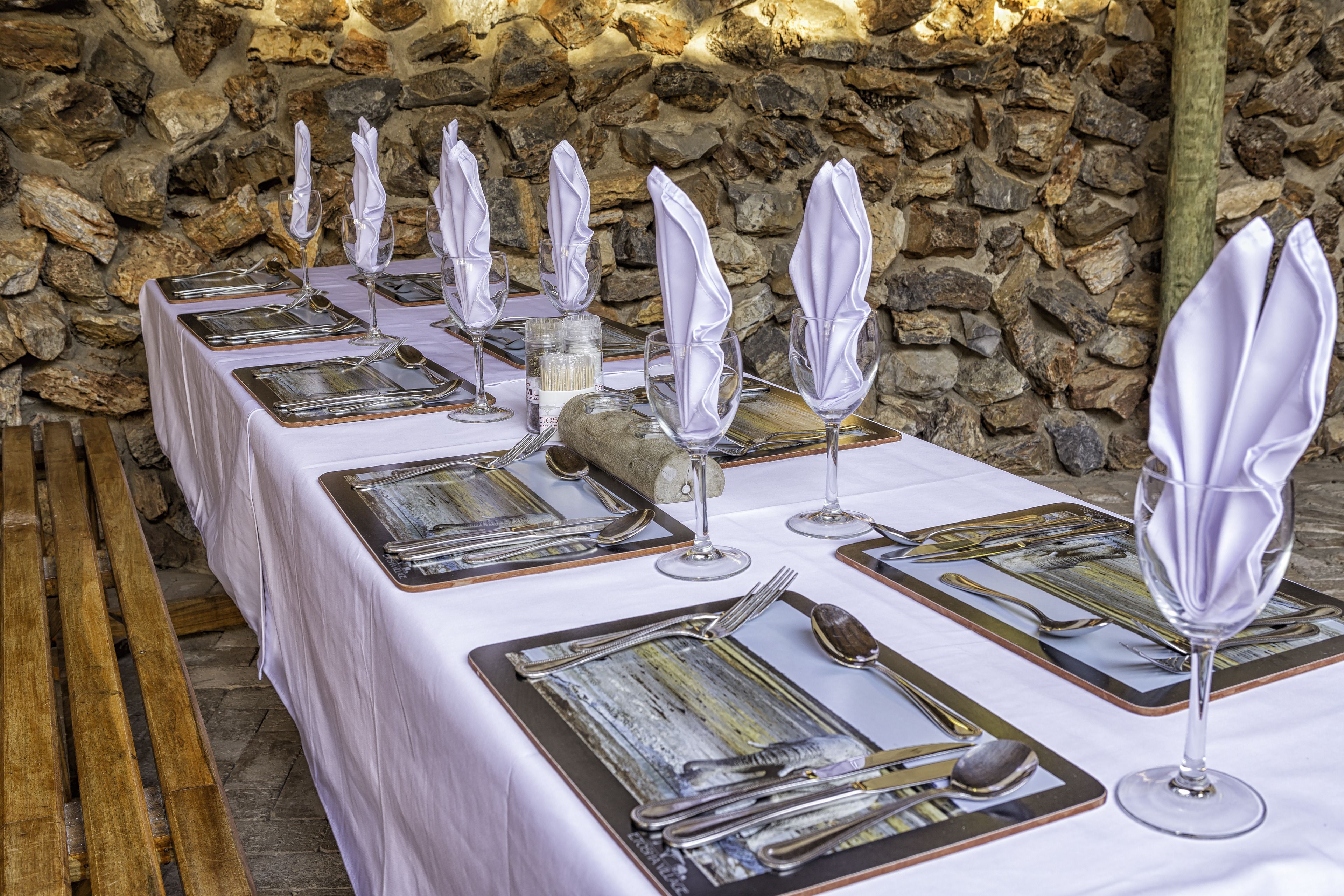 A long table set with white napkins, silverware, and wine glasses, against a stone wall backdrop.