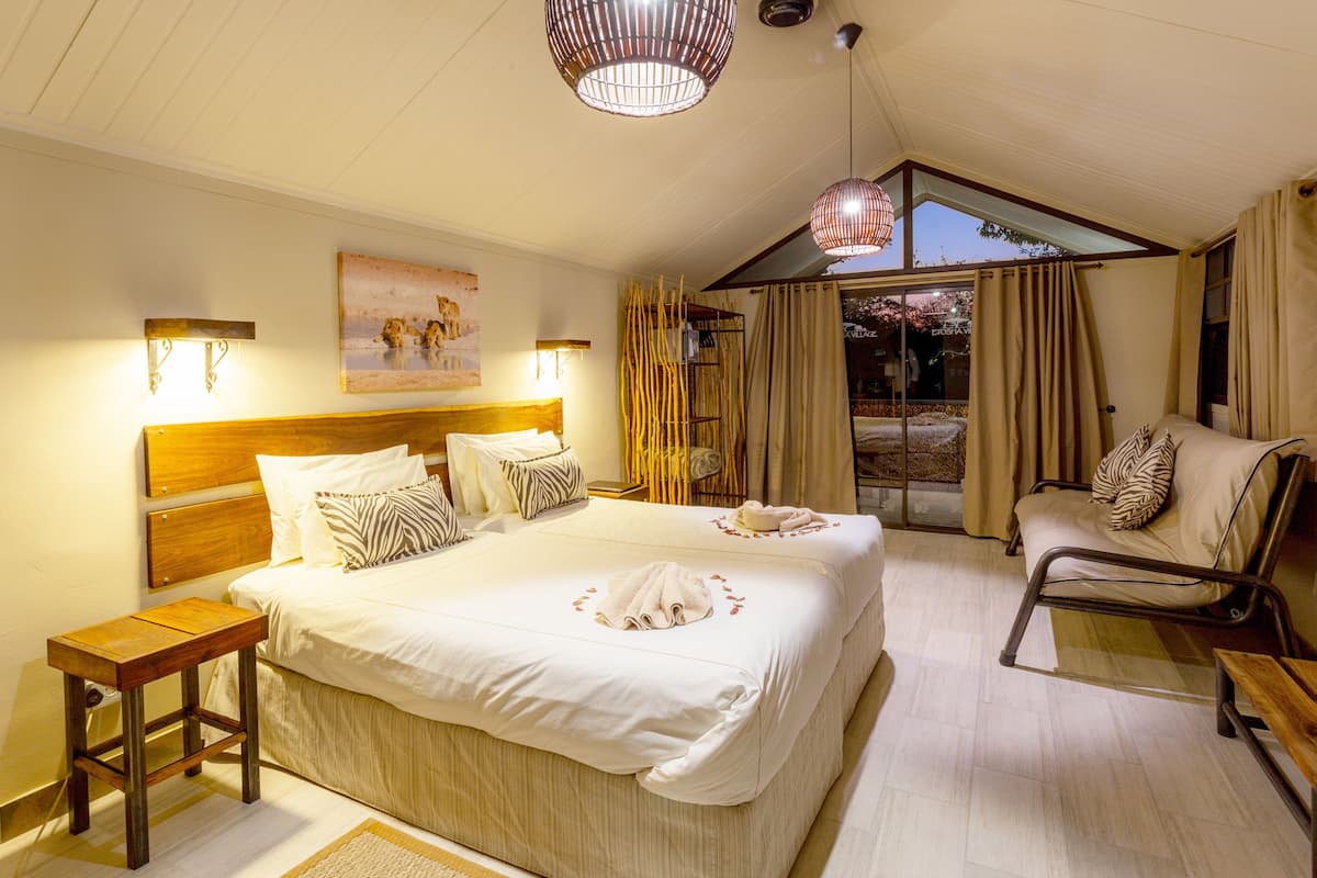 Un dormitorio amplio con una cama grande, una mesita de noche de madera, una zona de estar con una silla y vistas al exterior a través de un ventanal.