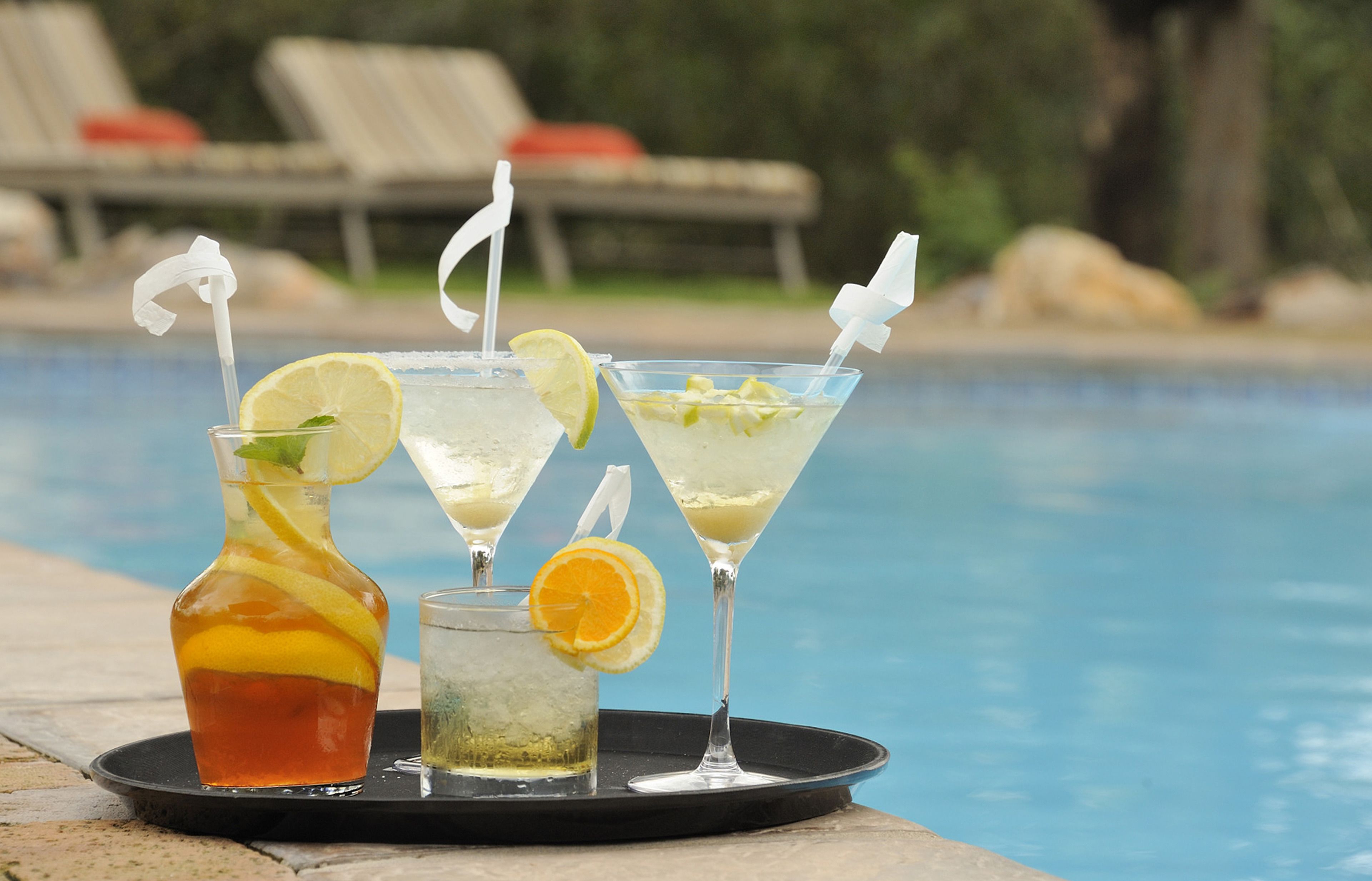 Una bandeja con tres bebidas cerca de una piscina, con sillones de descanso al fondo.