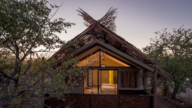 A rustic cabin with a thatched roof, a glass door entrance, and a stone foundation. - Etosha Village (Okaukuejo)