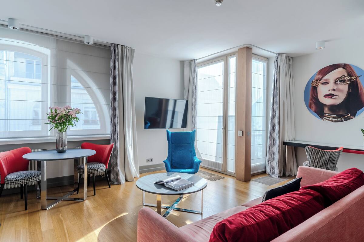 A modern living room with a round table, red and black patterned chairs, a blue armchair, and a red sofa. A flat-screen TV is mounted on the wall, and a large window provides natural light.