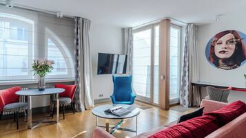 Een moderne woonkamer met een ronde tafel, stoelen met een rood-zwart patroon, een blauwe fauteuil en een rode bank. Een flatscreen televisie is aan de muur bevestigd, en een groot raam zorgt voor natuurlijk licht.