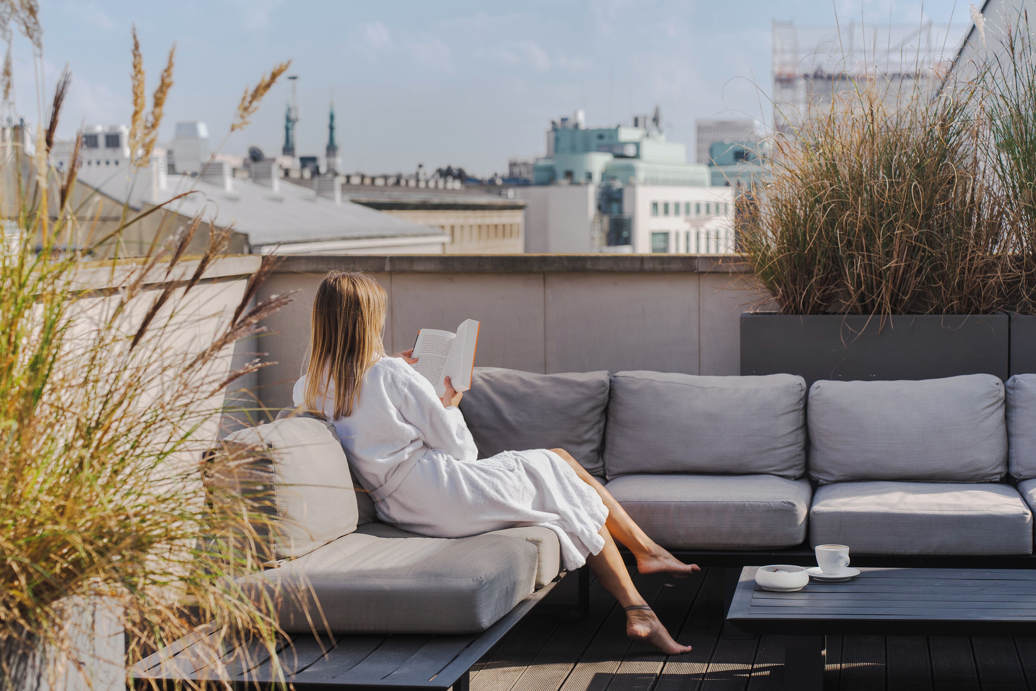 Una persona sentada en un sofá gris leyendo un libro en una terraza en la azotea, con vistas a edificios y una taza sobre una mesa.