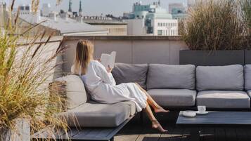 Una persona sentada en un sofá gris leyendo un libro en una terraza en la azotea, con vistas a edificios y una taza sobre una mesa.