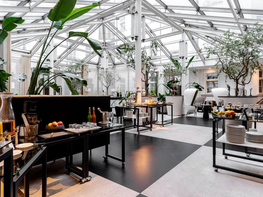 Amplio espacio interior con techo alto y paneles de vidrio, que incluye una mesa central con cafetera, rodeada de sillas y una mesa de buffet con comida y bebidas.