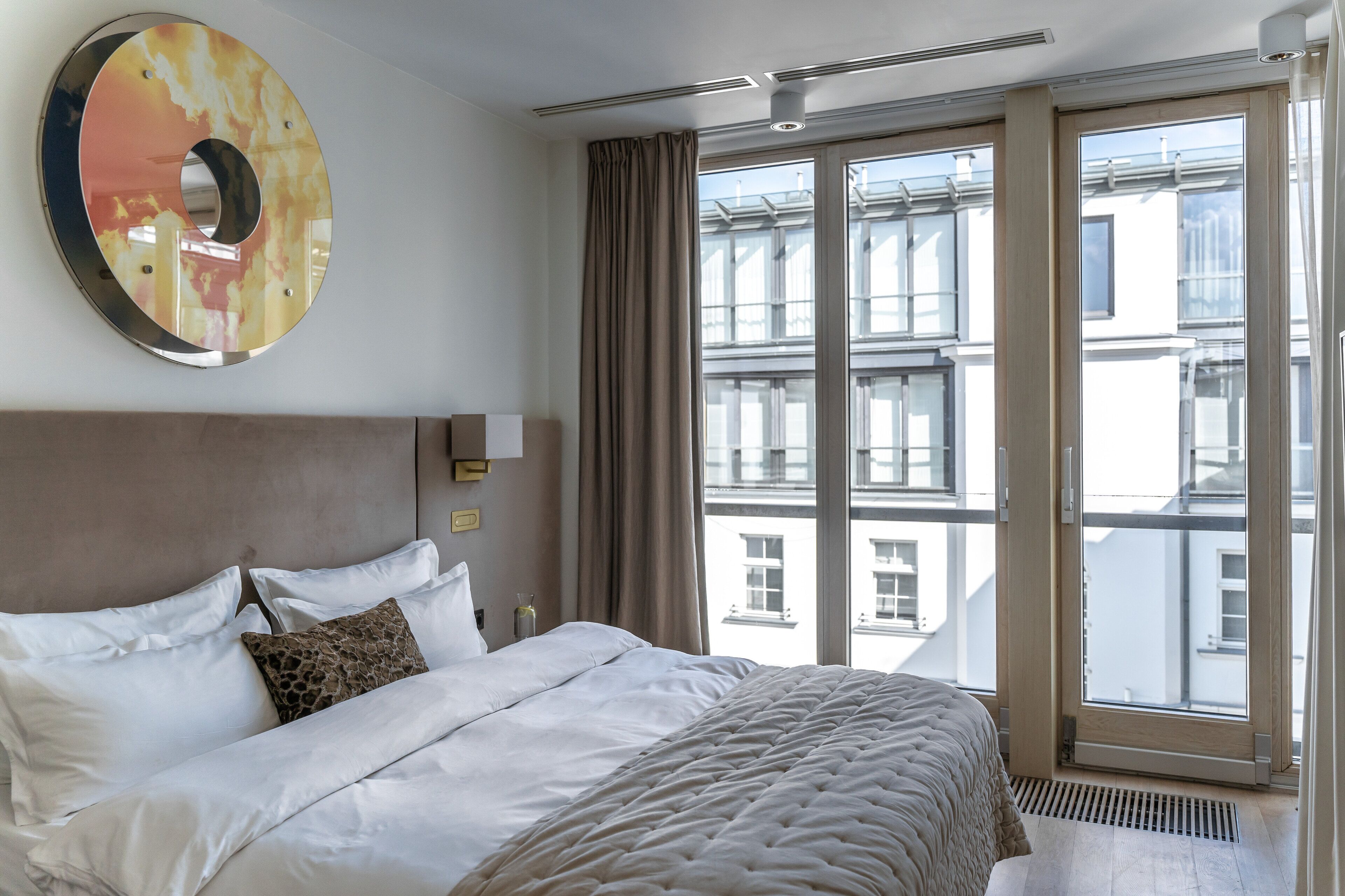 A hotel room with a large bed, a modern headboard, a large window with curtains, and a view of buildings outside.