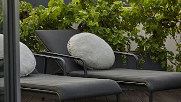 A row of black lounge chairs with grey cushions, positioned on a wooden deck, with a backdrop of green foliage.