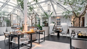 A spacious indoor area with a high ceiling and glass walls, featuring a dining setup with tables and chairs, surrounded by potted plants and modern decor.