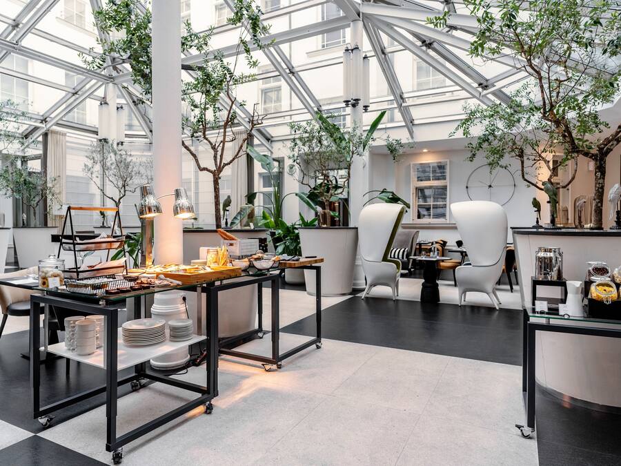 A spacious indoor area with a high ceiling and glass walls, featuring a dining setup with tables and chairs, surrounded by potted plants and modern decor.