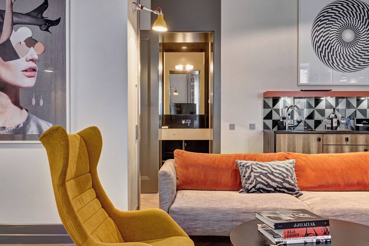 A modern living room with a yellow armchair, a sofa with orange cushions, a coffee table with books, and a kitchen area with a geometric backsplash.