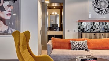 Un salón moderno con un sillón amarillo, un sofá con cojines naranja, una mesa de centro con libros y una zona de cocina con salpicadero geométrico.
