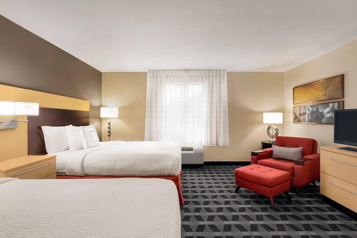 Ein Hotelzimmer mit zwei Betten, einem roten Sessel, einem Fernseher und einem großen Fenster mit Vorhängen.