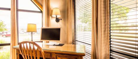 A wooden desk with a computer monitor, a lamp, and a chair in a room with large windows and blinds.
