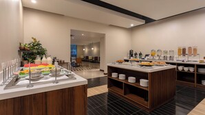 A buffet setup with various food items and beverages, a seating area in the background, and a modern interior design. - Four Points by Sheraton San Jose Sabana (San José)