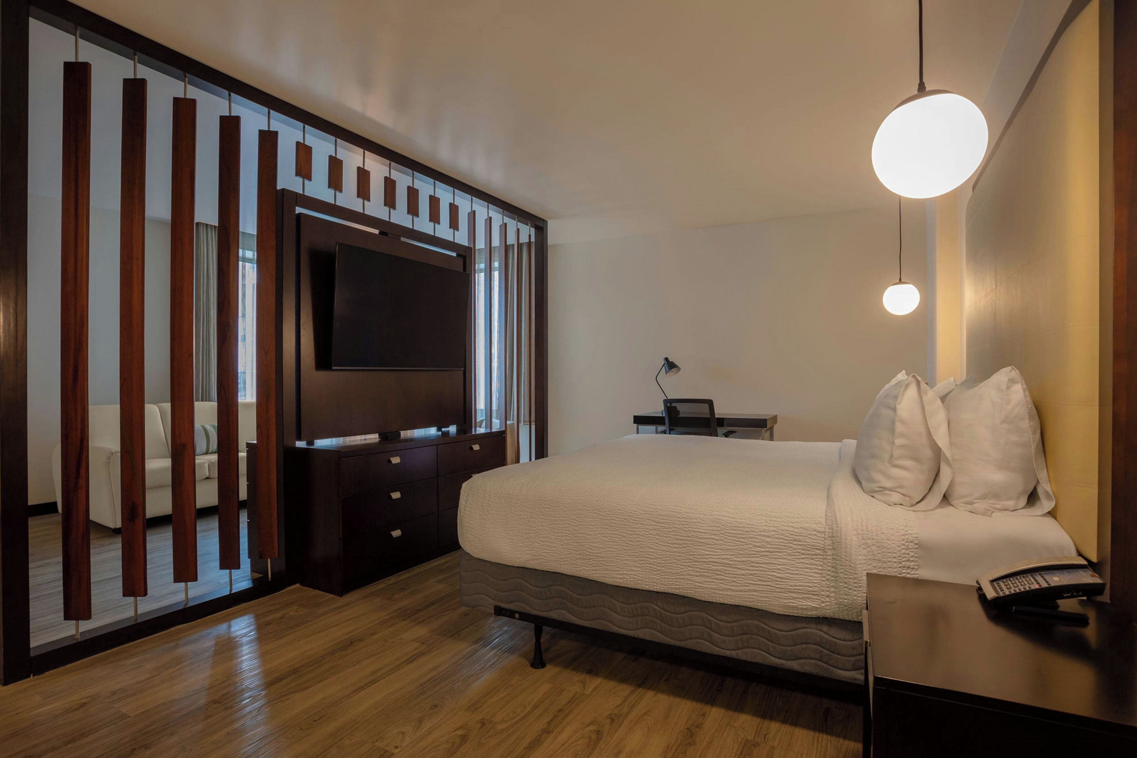 A modern bedroom with a large bed, a TV mounted on a wooden partition, a desk with a lamp, and a telephone.